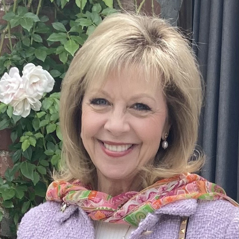 Tracy Lindsey smiling in a lavender jacket and floral scarf beside climbing roses.