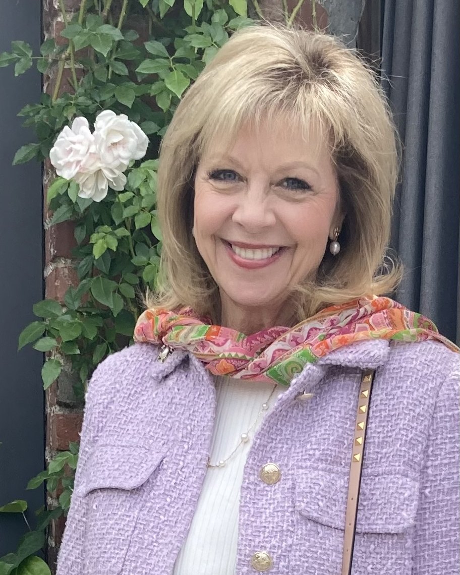 Tracy Lindsey smiling in a lavender jacket and floral scarf beside climbing roses.